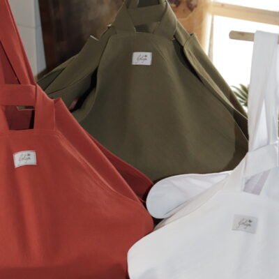 Close-up shot of the three colors in the Artisanal Casserole Carrier Kit: terracotta orange, olive green, and classic white, sitting on a wooden counter with the Goligoi Maison tag visible. The carriers are bunched up, showing the strong handles for safe transport.